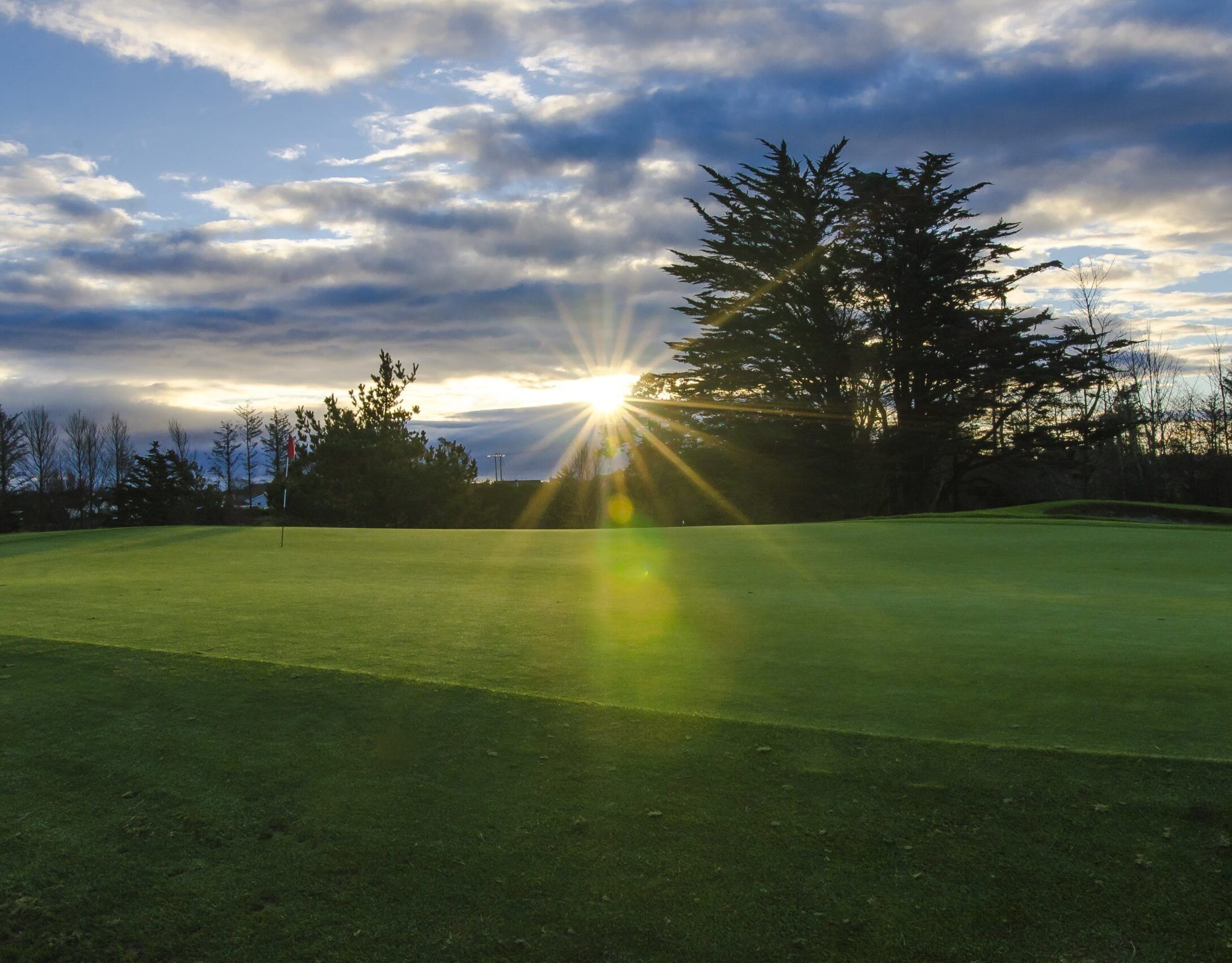 Un campo de golf con un mástil de bandera, rodeado de árboles y una puesta de sol al fondo.