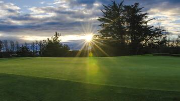 Un campo de golf con un mástil de bandera, rodeado de árboles y una puesta de sol al fondo.