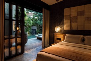 A bedroom with a large bed, a nightstand, a wall-mounted lamp, and a sliding glass door leading to an outdoor area with a pool and greenery.