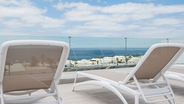 Dos sillones blancos con cojines beige en un balcón con vistas despejadas al mar y edificios al fondo.