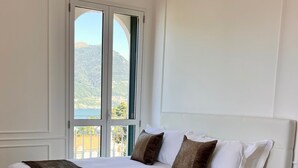 Ein großes Bett mit weißer Bettwäsche und einer braunen Decke, platziert in einem Zimmer mit Holzböden und Blick auf eine gebirgige Landschaft durch ein großes Fenster.
