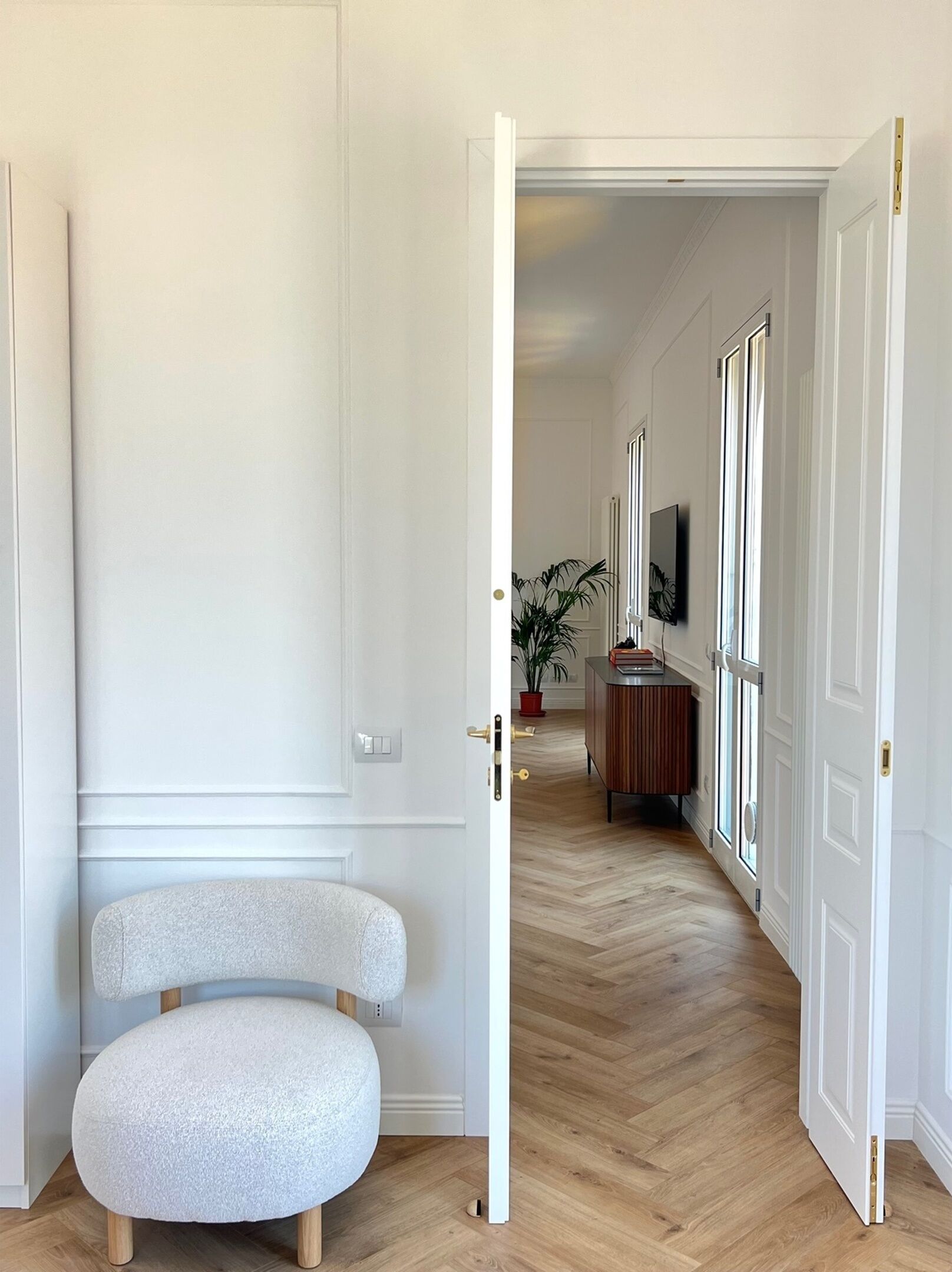 Um corredor com uma porta branca, um banquinho redondo, uma planta em vaso e um armário de madeira.