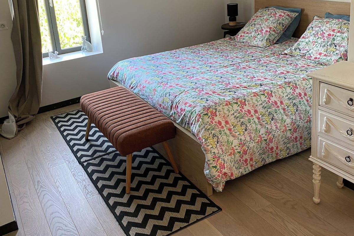 Un dormitorio con cama, un banco, un tocador, una mesita de noche, una ventana con cortinas y dos cuadros en la pared.