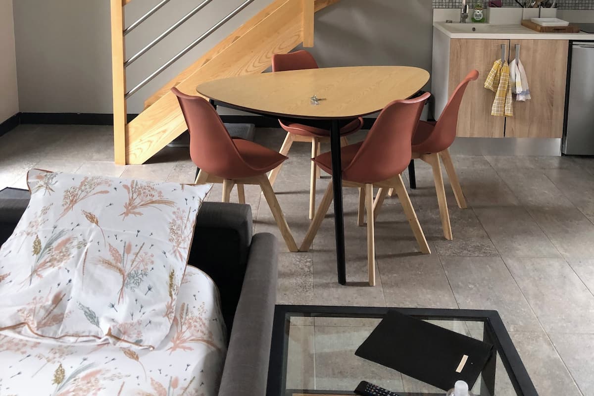 Una sala de estar moderna con un sofá, una mesa redonda y sillas. Una escalera con barandilla de madera conduce a un nivel superior.