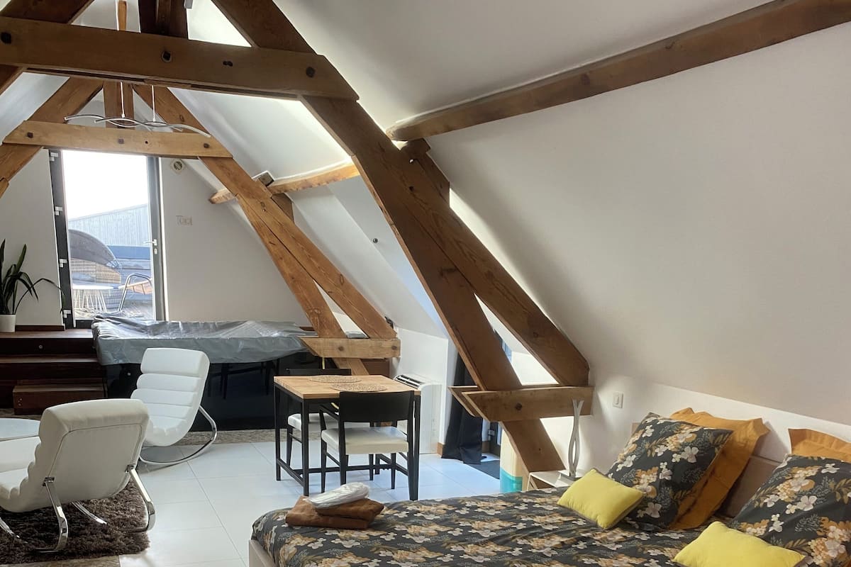 Un dormitorio con techo inclinado, una cama con ropa de cama floral, una silla de madera, una mesita y vistas al exterior.