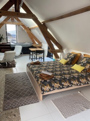 Una camera da letto con soffitto spiovente, un letto con lenzuola a motivi floreali, una sedia di legno, un piccolo tavolo e vista sull'esterno.
