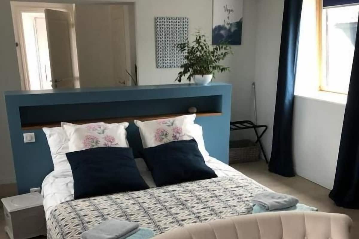 Un dormitorio con cama, una silla, una mesita de noche, una planta y una ventana.