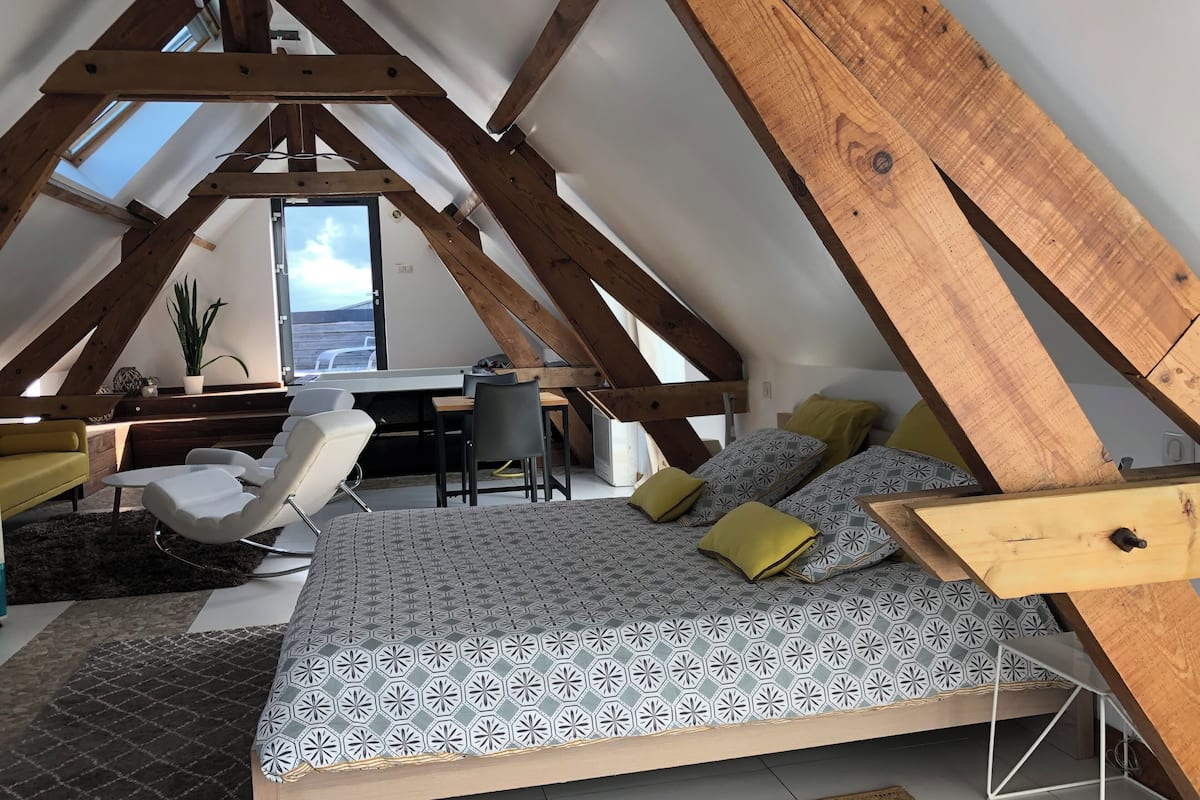 Un dormitorio tipo loft con techo inclinado, una cama con ropa de cama estampada, un escritorio con una silla y una planta en maceta.