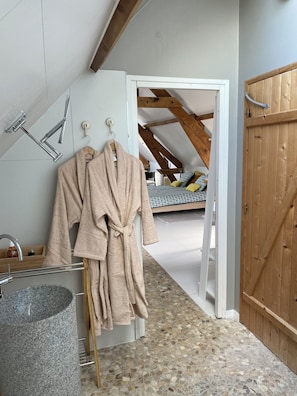 Een badkamer met een stenen wastafel, een kledingrek met twee gewaden en een houten deur.