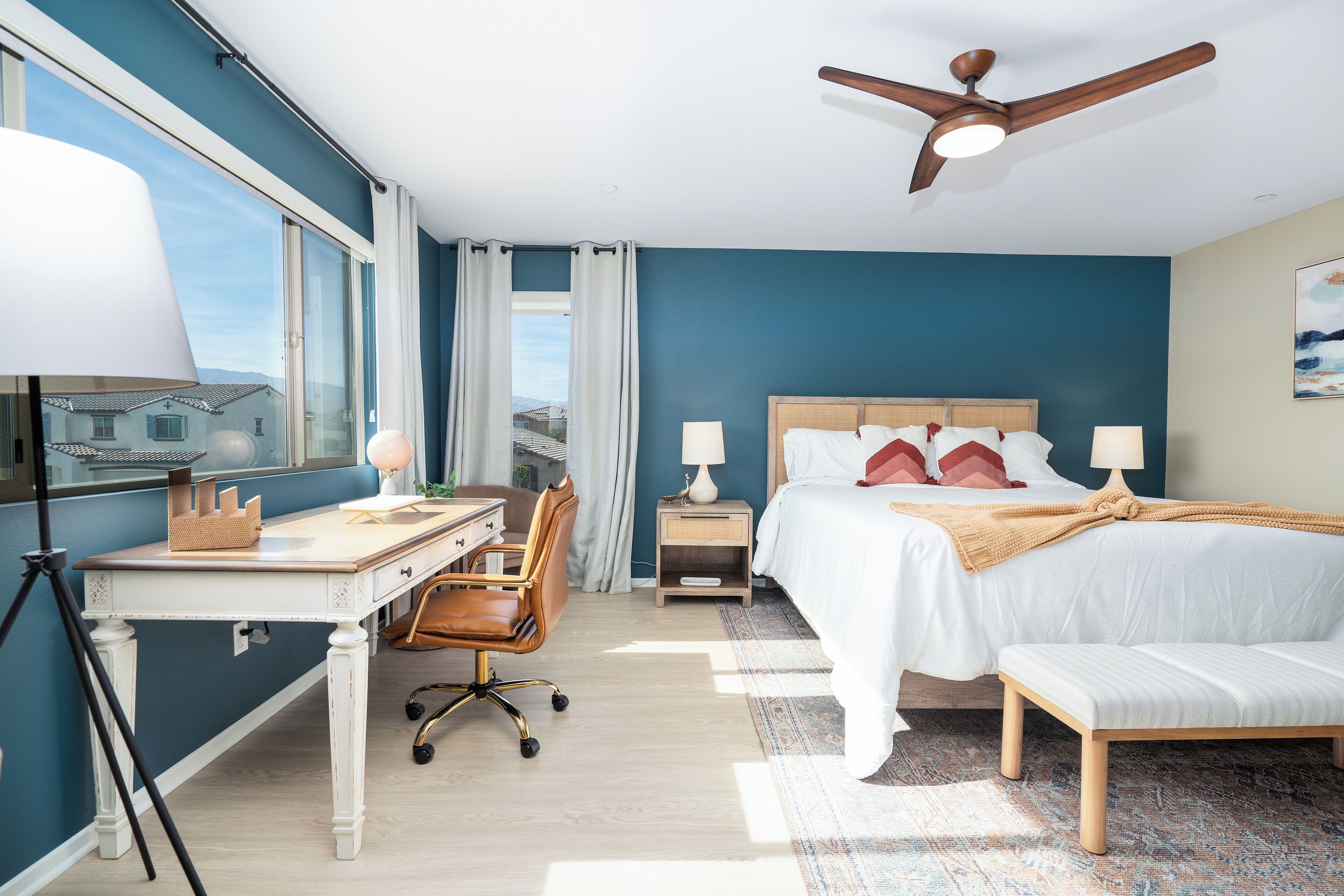 A hotel room with a desk, chair, bed, bedside tables, lamps, and a ceiling fan.