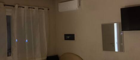 A room with a beige sofa, a white desk, a hanging pendant light, a window with curtains, and an air conditioning unit.
