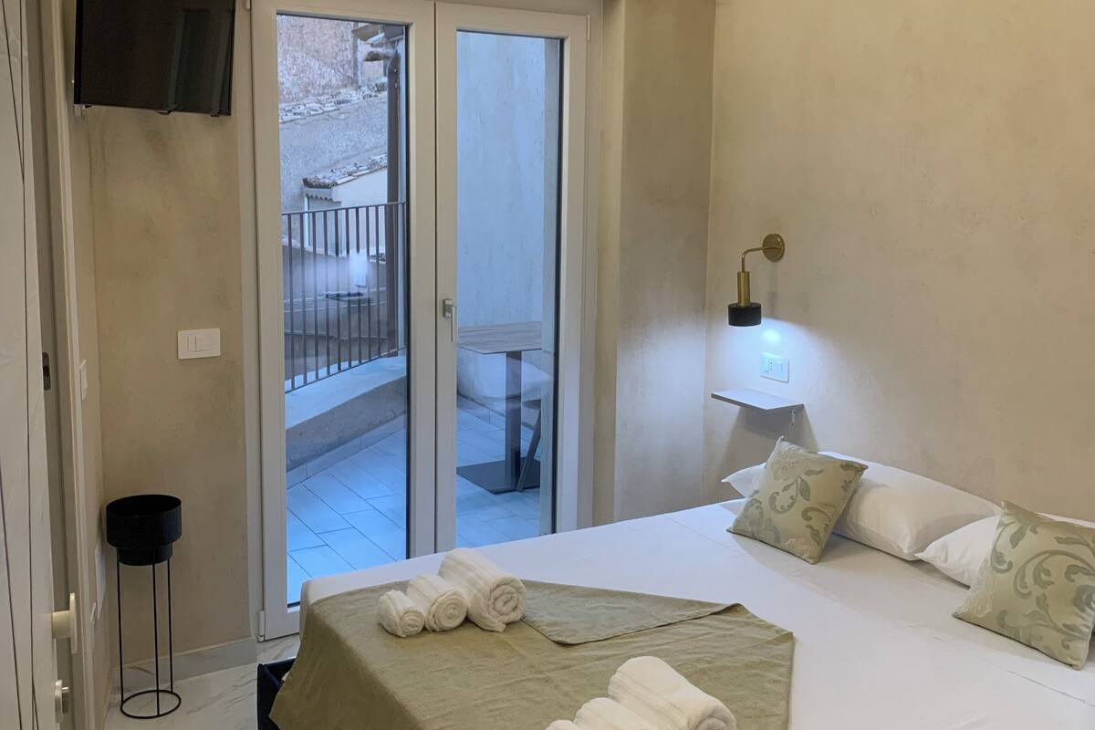 Une chambre avec un lit, une table de chevet avec une lampe, une porte-fenêtre coulissante donnant sur un balcon extérieur et un téléviseur fixé au mur.