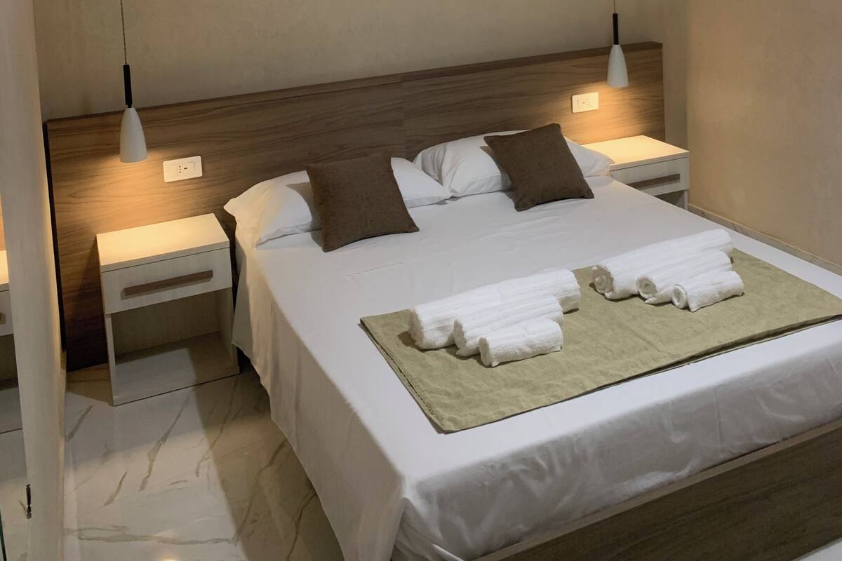 Une chambre moderne avec une tête de lit en bois, un lit recouvert de draps blancs et agrémenté d’oreillers marron, deux tables de chevet avec des lampes et un sol en marbre.