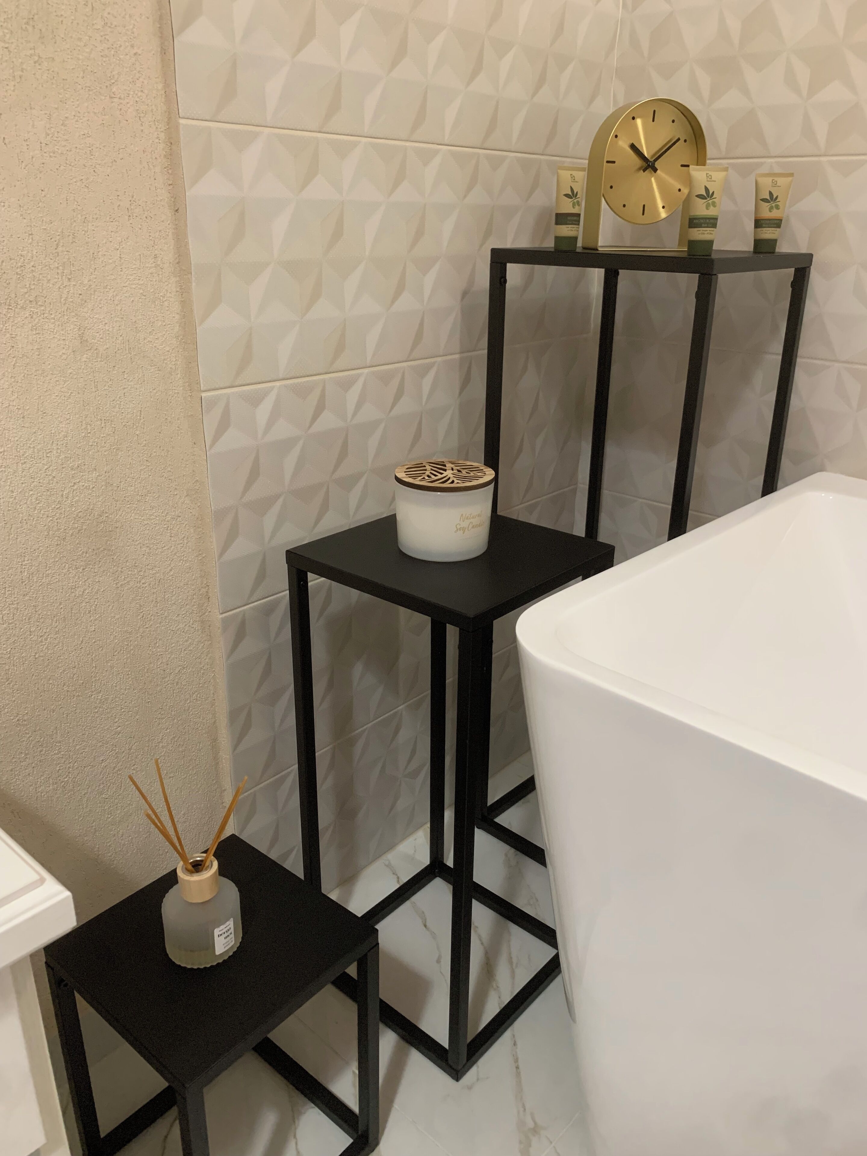Une salle de bain avec une baignoire blanche, une étagère en métal noir avec une horloge et des articles de toilette, et une petite table sur laquelle se trouve un diffuseur de parfum à bâtonnets.