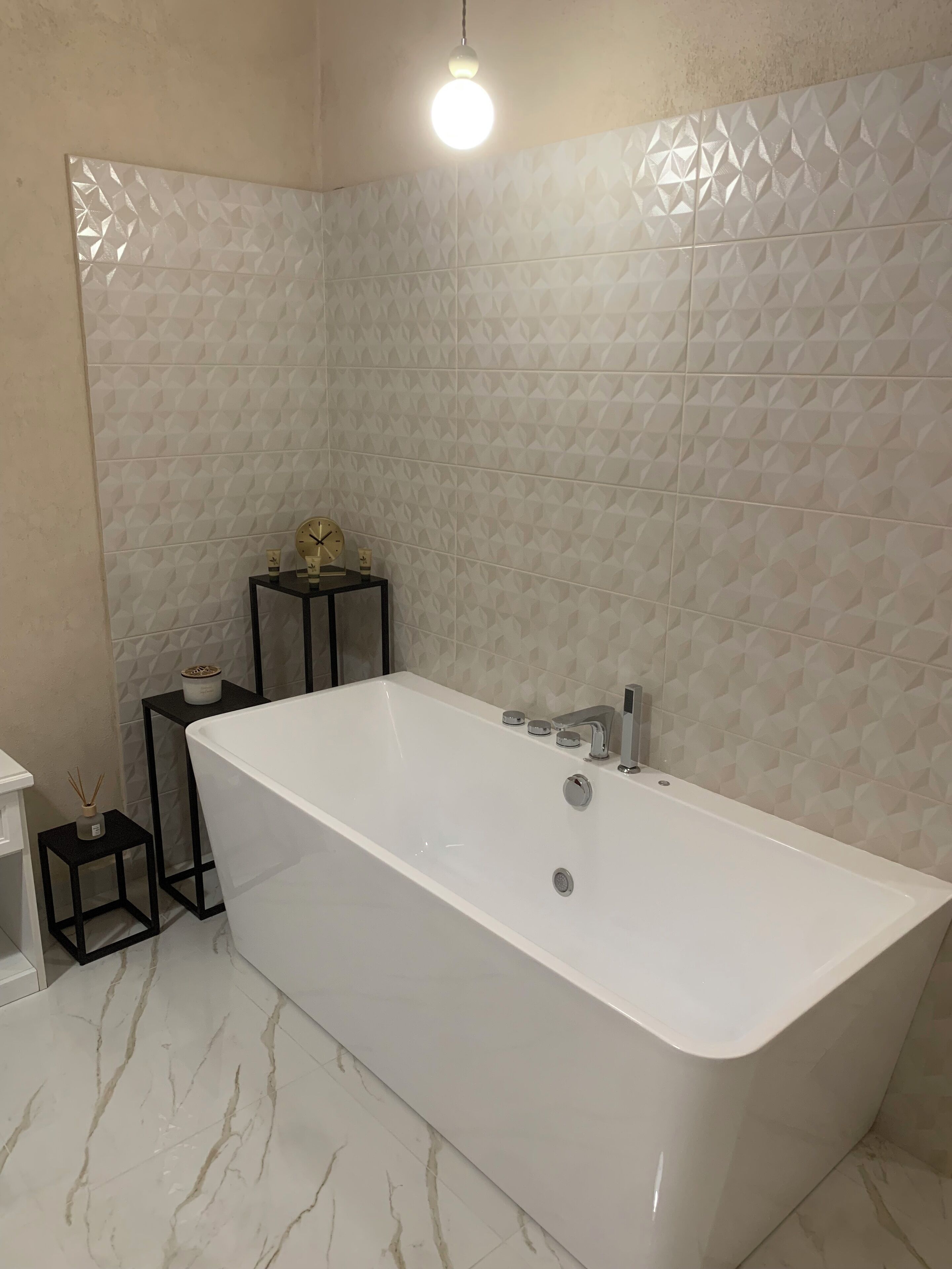 A modern bathroom with a large white bathtub, a small black side table with a clock and a candle, and a textured wall.