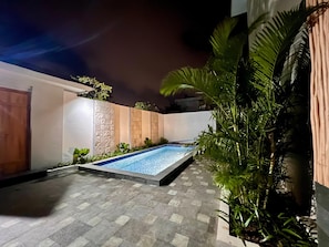 A rectangular swimming pool with a tiled deck, surrounded by a low wall with textured panels and plants.