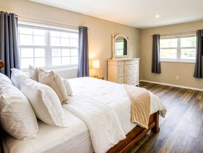 A bedroom with a large bed, a dresser, a mirror, and two windows with curtains.