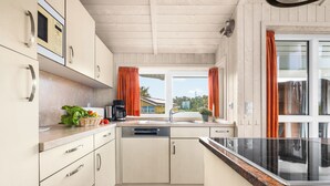 A modern kitchen with white cabinetry, a black countertop, and a window with orange curtains. - Möwenstübchen Ferien House in Grömitz: Sauna, A/C, 250 m to beach (Grömitz)