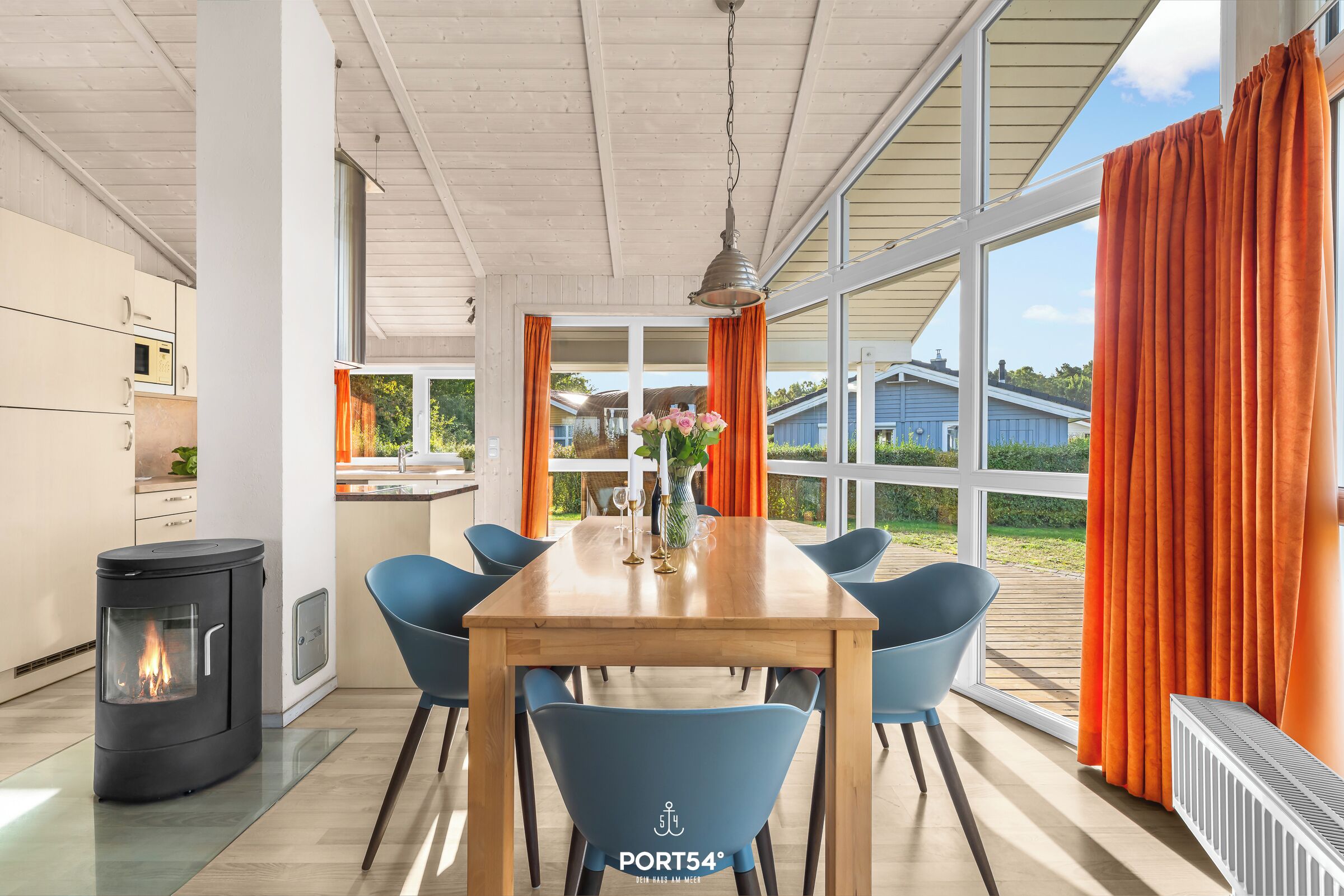 Eine moderne Küche mit Einbauherd, ein Essbereich mit Holztisch und blauen Stühlen sowie große Fenster mit orangefarbenen Vorhängen.