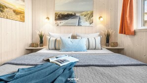 A bedroom with a wooden wall, a bed with striped pillows, a blue blanket, and a painting of a beach. - Möwenstübchen Ferien House in Grömitz: Sauna, A/C, 250 m to beach (Grömitz)