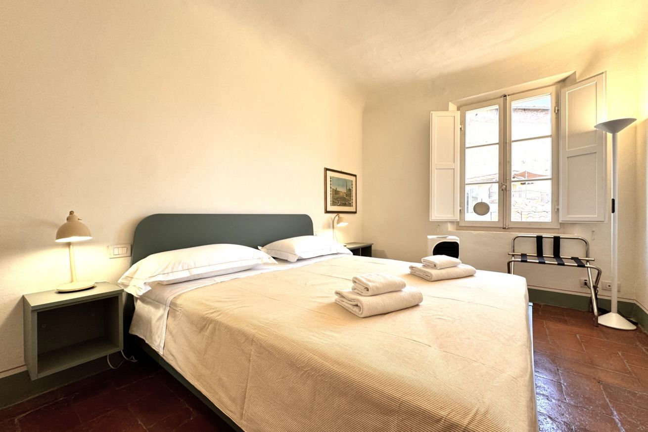 Un letto rifatto con lenzuola e asciugamani bianchi, una lampada da comodino e una finestra con persiane.