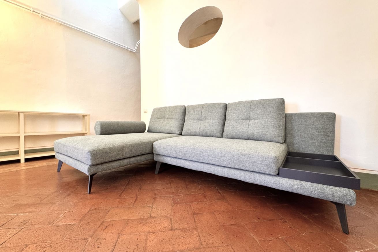 A grey sectional sofa with a black tray, a wooden bench, and a circular wall decoration in a room with a tiled floor.