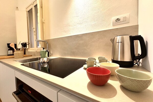 Una cocina moderna con una placa de inducción negra, un hervidor de acero inoxidable y varios cuencos y tazas sobre la encimera.