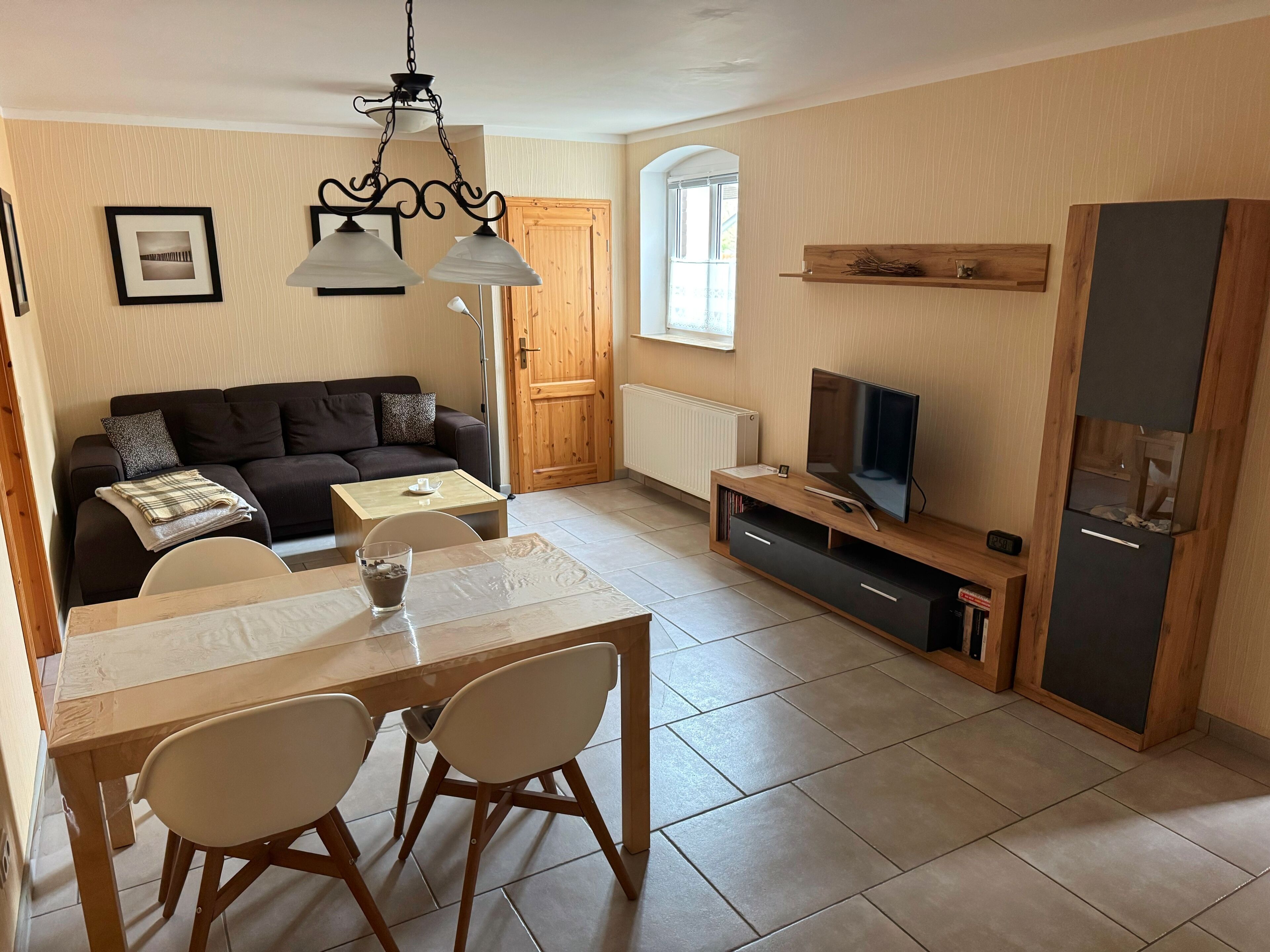 Ein Wohnzimmer mit Sofa, Fernseher, Holz-TV-Möbel, Esstisch mit Stühlen und einer Chandelüster.