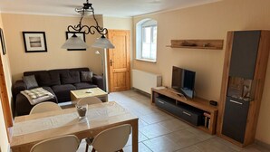 Un salon comprenant un canapé, un téléviseur, un meuble TV en bois, une table à manger avec des chaises et un lustre.
