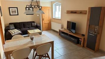 Ein Wohnzimmer mit Sofa, Fernseher, Holz-TV-Möbel, Esstisch mit Stühlen und einer Chandelüster.