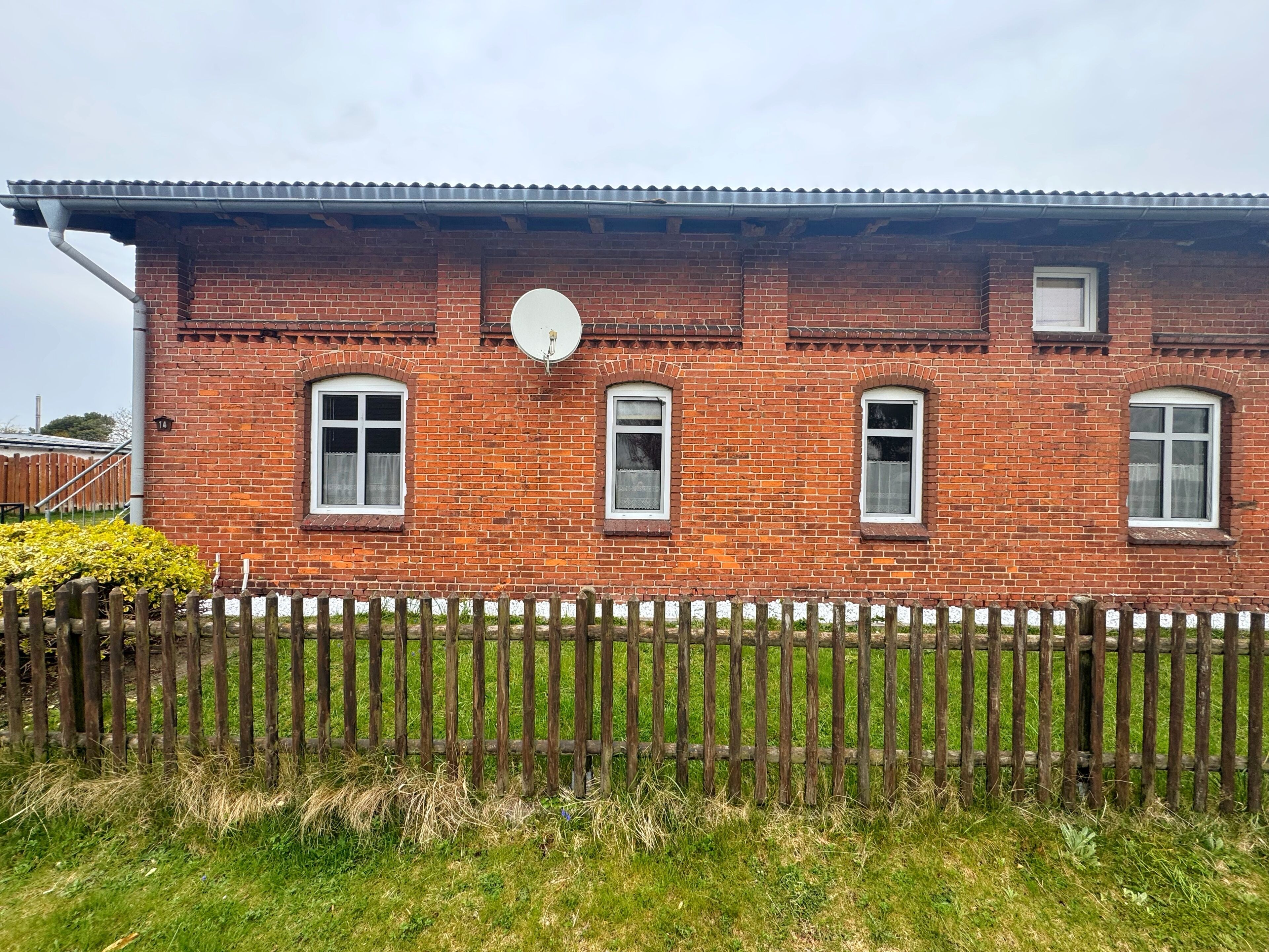 Ein Backsteinhaus mit Satellitenschüssel, drei Fenstern und einem Holzzaun.