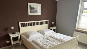 Une chambre avec un lit blanc, deux tables de chevet, une commode, une fenêtre donnant sur un paysage agréable et un tableau encadré au mur.