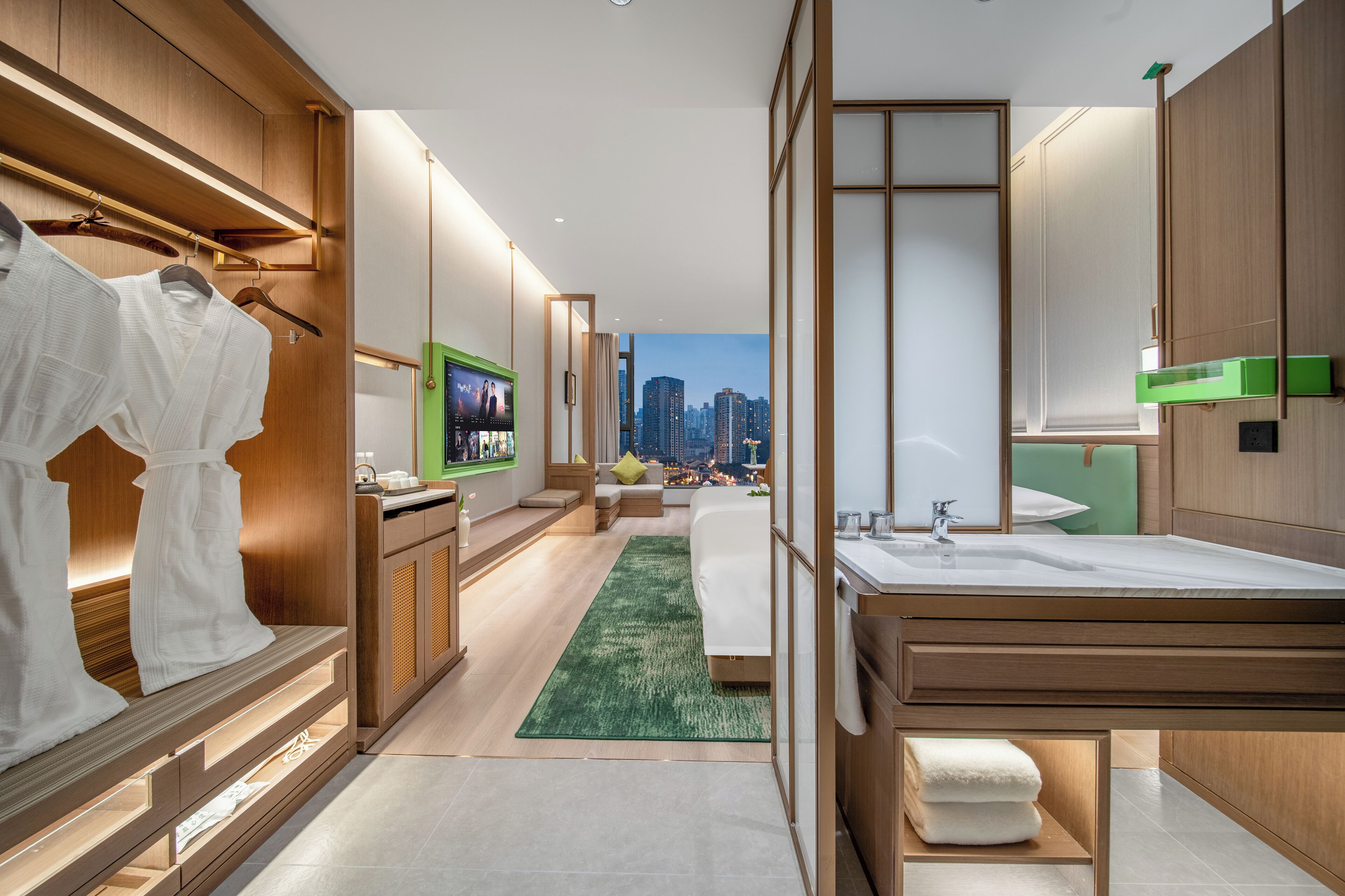 A modern hotel room with a city view, a walk-in closet, a large bed, and a bathroom with a green accent wall.