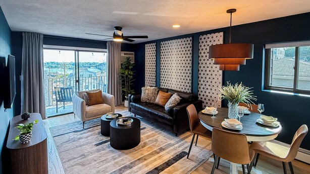 A modern living room with a sofa, coffee table, dining table, and chairs. There is a ceiling fan, a TV mounted on the wall, and a view of the city through large windows. - City Views~Close 2 Salem~Balcony (Beverly)