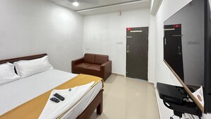 A hotel room with a bed, a brown armchair, a TV, and a door with a number sign.