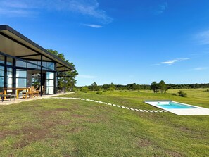 Una casa moderna con un amplio ventanal de vidrio, mobiliario de exterior y una piscina.