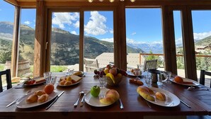 Ein Holztisch gedeckt mit Tellern, Brot, Obst und Gläsern, mit Blick auf eine Berglandschaft.
