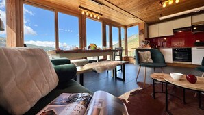 Ein gemütliches Wohnzimmer mit einer Couch, einem Tisch mit einer Zeitschrift und Blick auf eine Berglandschaft durch große Fenster.