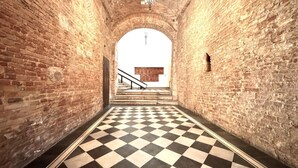 A corridor with checkered flooring and brick walls, leading to a staircase with a wooden handrail. - Casa Bruna- Via banchi di sotto (Siena)