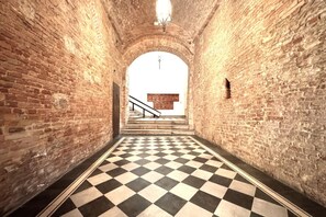 A corridor with checkered flooring and brick walls, leading to a staircase with a wooden handrail. - Casa Bruna - Via Banchi di Sotto (Siena)