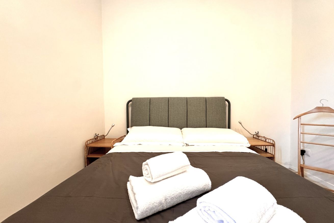 Un letto rifatto con una coperta marrone scuro e cuscini bianchi, un comodino in legno con una lampada e una sedia in legno con un appendiabiti.