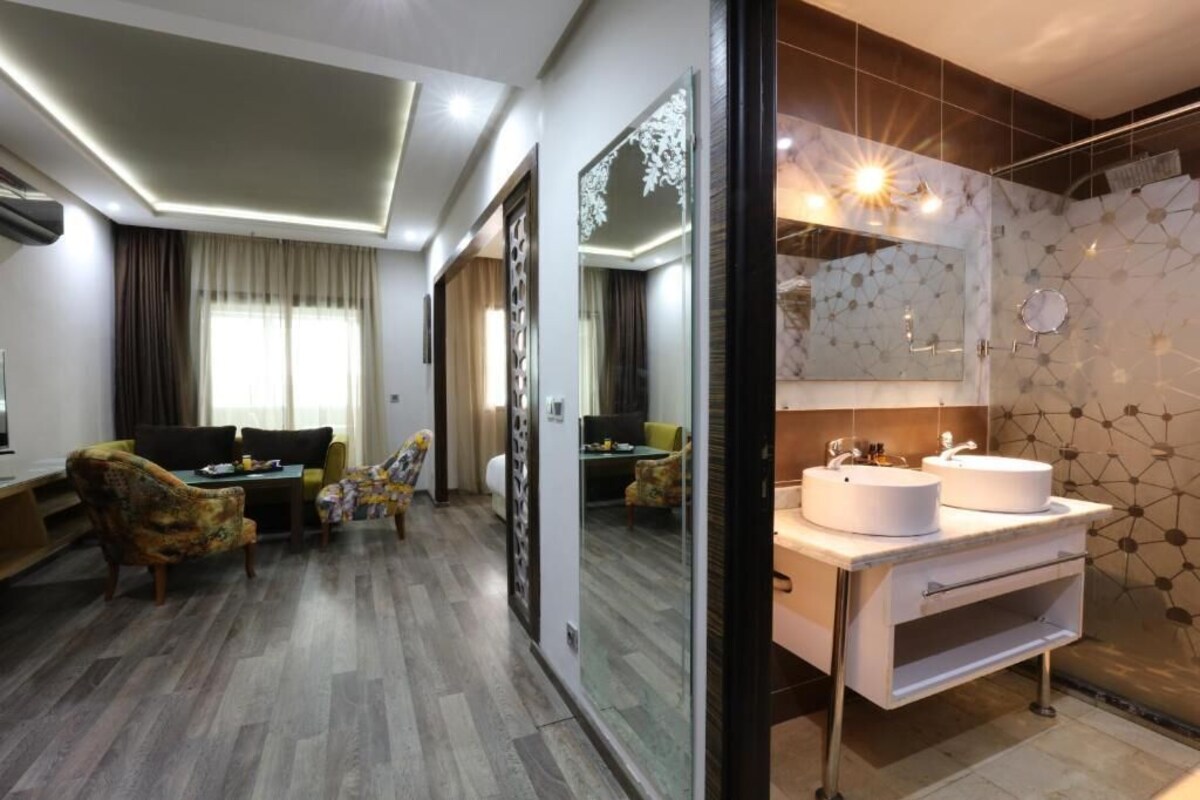 A modern hotel room with a living area, a large mirror, and a bathroom with two sinks.