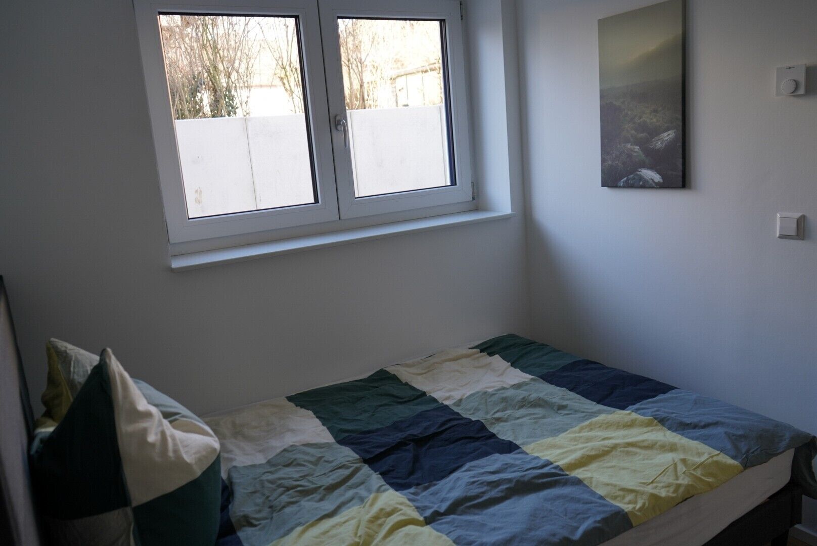 Une petite chambre avec un lit simple, une fenêtre, un tableau au mur et un thermostat.