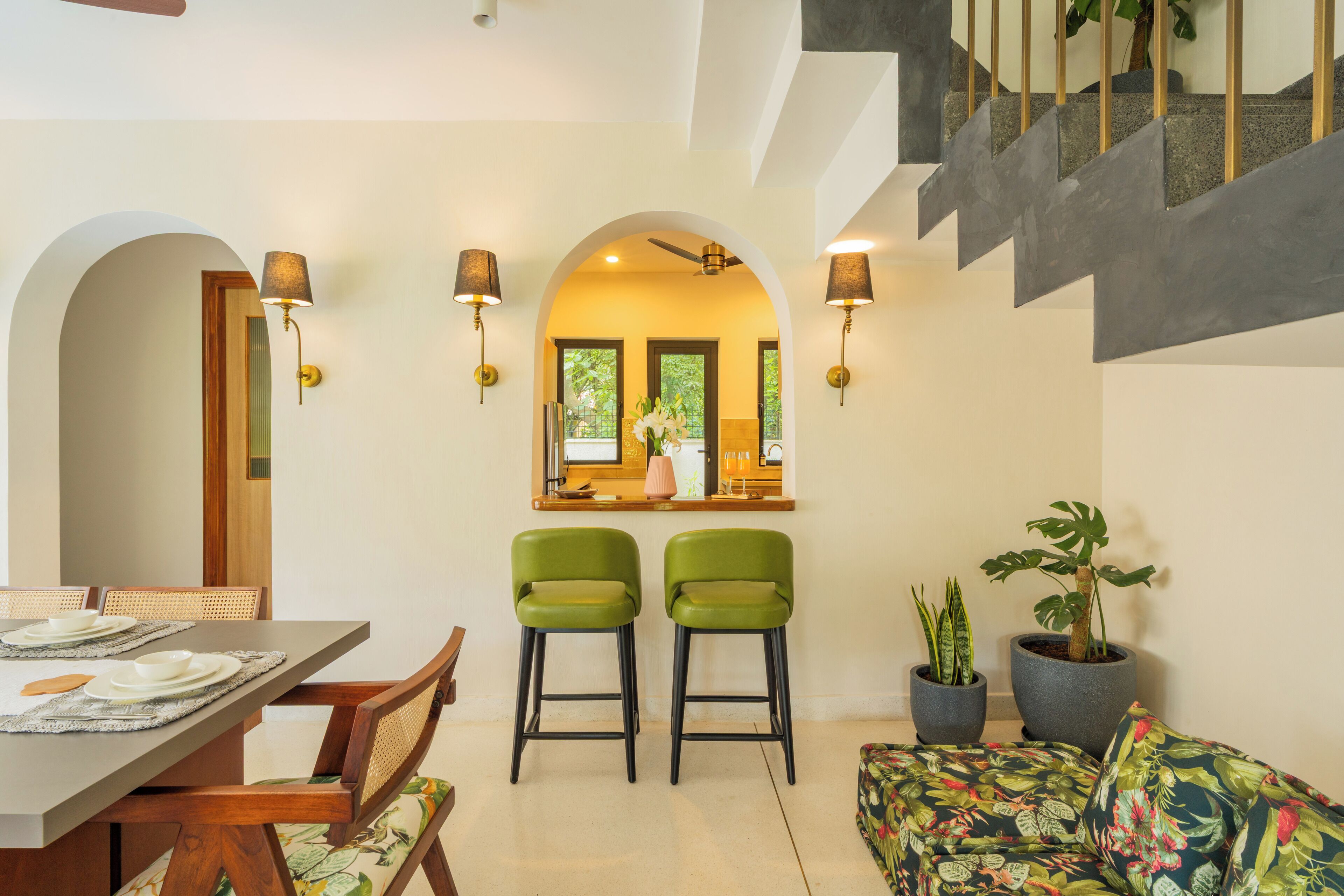 A dining area with a table set for two, two green chairs, a sofa with a floral pattern, and two potted plants.