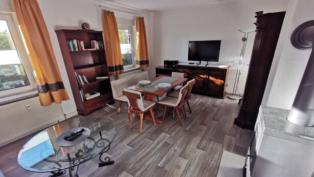 A modern living room with a glass coffee table, wooden dining table, and chairs. There is a bookshelf, a flat-screen TV, and a fireplace. - Cozy vacation apartment on the outskirts of Pirna in Saxon Switzerland (Pirna)