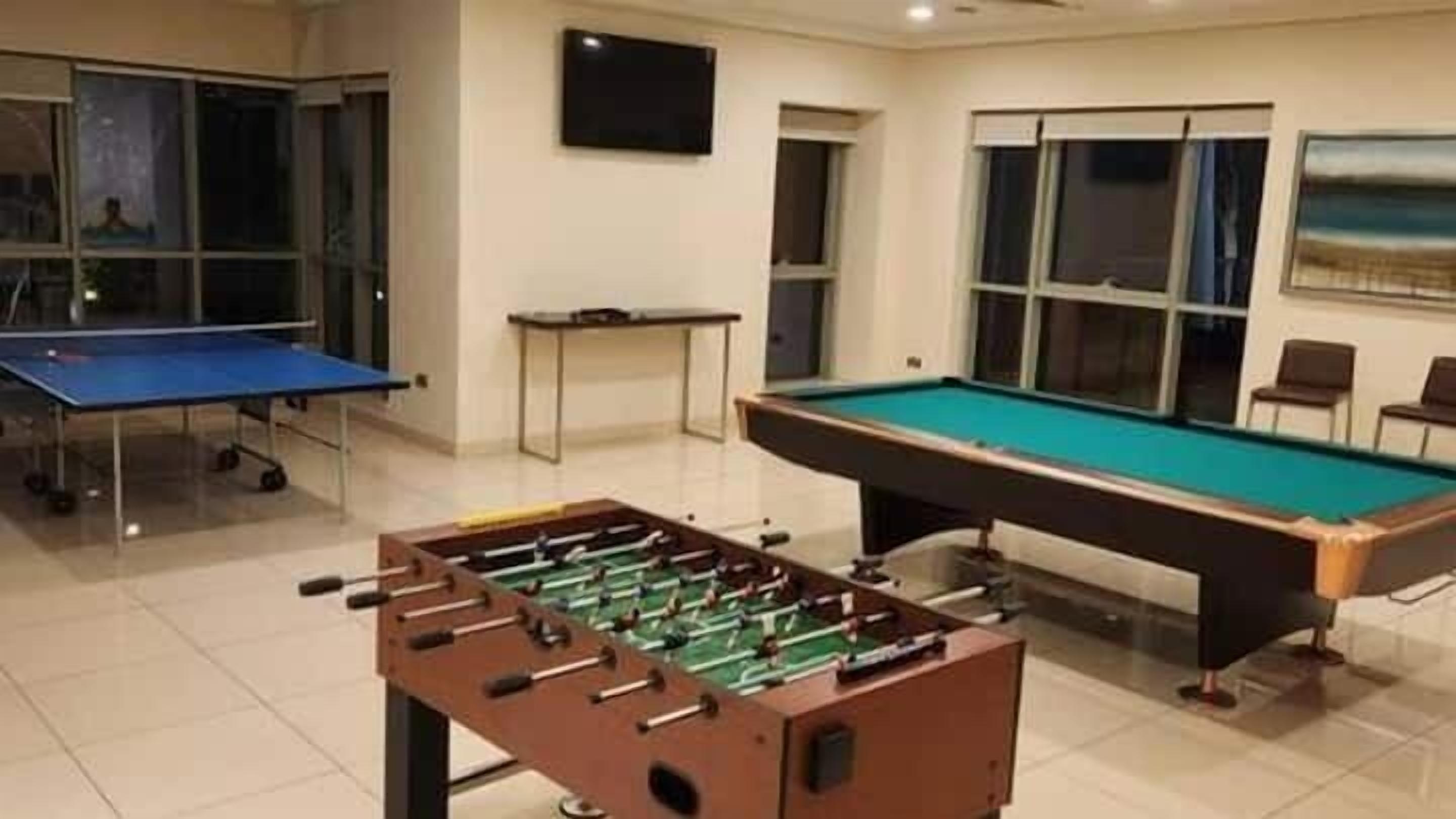 Ein Billardtisch, ein Kicker und ein Pong-Tisch in einem Raum mit Fenstern und einem Fernseher.