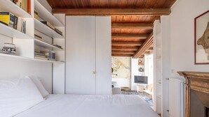 A bedroom with a large bed, white walls, wooden ceiling beams, a bookshelf, and a fireplace.