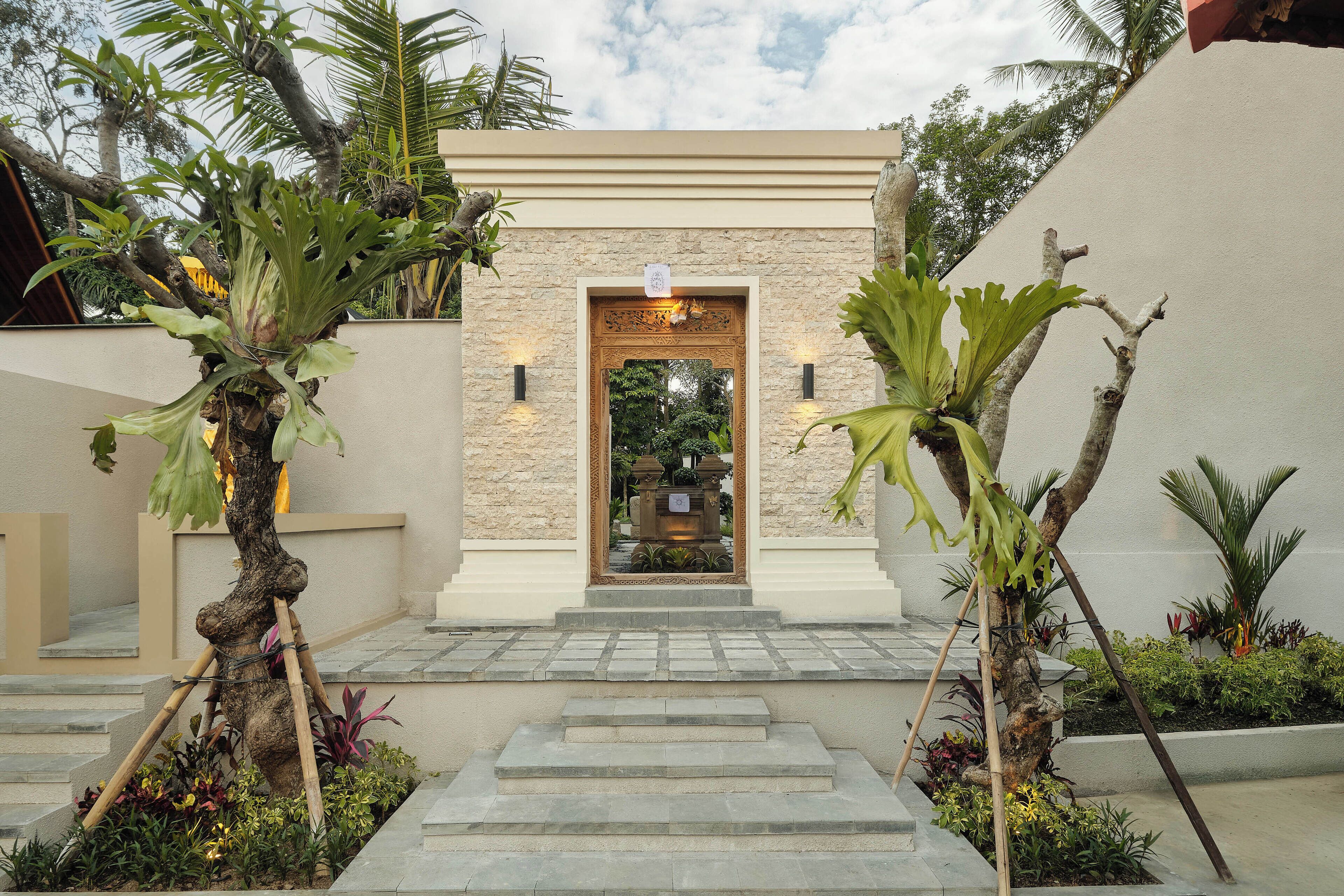 A building entrance with a stone facade, a wooden door, and a small garden area with plants and steps leading up to it.
