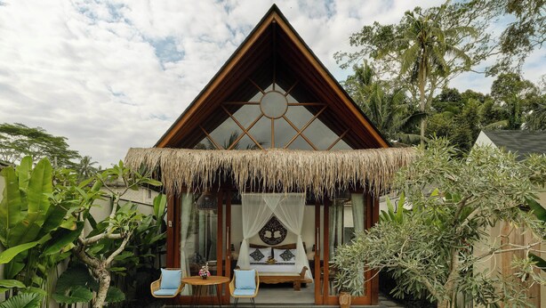 A triangular-roofed building with a thatched roof entrance, a bed with a canopy, and a small outdoor seating area with chairs. - Changi Jungle Ubud Villas (Ubud)