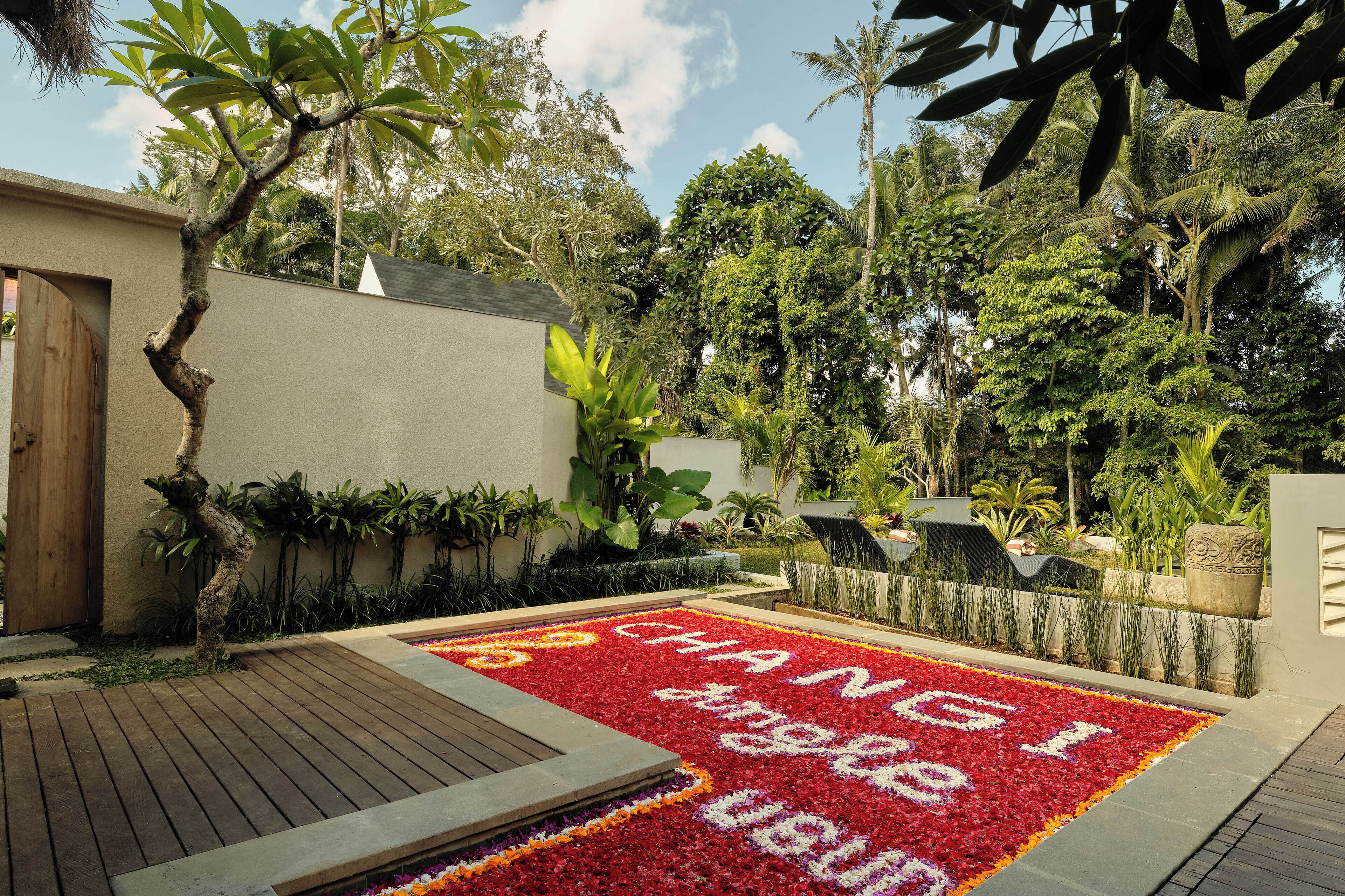 A garden with a wooden deck, a flower bed with red and white flowers spelling out "CHANGI," and various tropical plants.